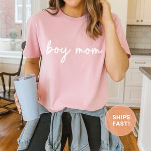 K&ouml;nnte beinhalten: Ein hellrosa T-Shirt mit den Worten "boy mom" in wei&szlig;er Schrift. Das Shirt hat kurze &Auml;rmel und scheint aus einem weichen Material zu sein. Ein hellblauer Becher wird in der linken Hand gehalten.