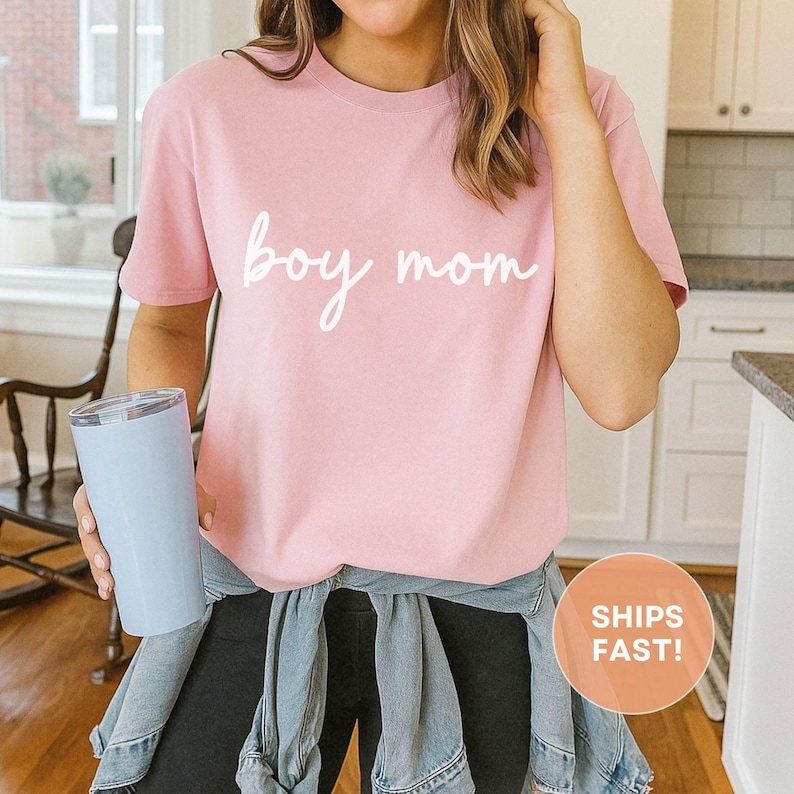 K&ouml;nnte beinhalten: Ein hellrosa T-Shirt mit den Worten "boy mom" in wei&szlig;er Schrift. Das Shirt hat kurze &Auml;rmel und scheint aus einem weichen Material zu sein. Ein hellblauer Becher wird in der linken Hand gehalten.