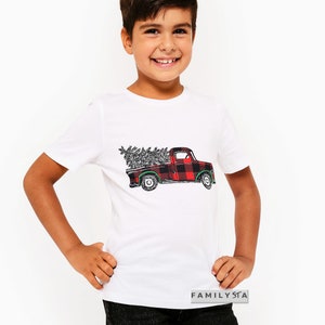 May include: White t-shirt with a red and black plaid pickup truck carrying a Christmas tree. The truck has a green steering wheel and green wheels. The text "FAMILYSTA" is on the bottom right corner of the shirt.