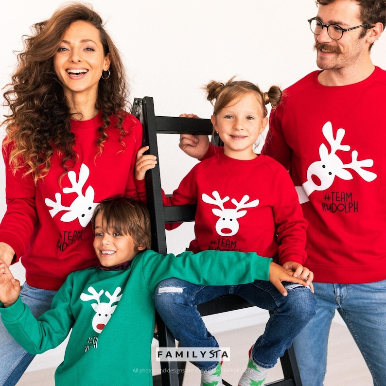 May include: A family wearing matching red and green Christmas sweaters with a white reindeer graphic. The sweaters have the text "#Team Rudolph" and "#Team". The family is smiling and posing for a photo.