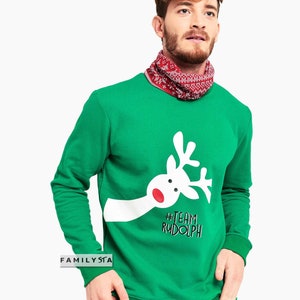 May include: A green sweatshirt featuring a white reindeer graphic with a red nose and the text "#TEAM RUDOLPH". The sweatshirt is paired with a red and white patterned neck gaiter, perfect for the holiday season.