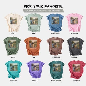 K&ouml;nnte beinhalten: Eine Auswahl an Comfort Colors T-Shirts in verschiedenen Farbt&ouml;nen, darunter Elfenbein, Bay, Blue Jean und Blossom. Jedes Shirt zeigt eine Grafik eines Hundes in einem Stuhl mit dem Text "I live here now".