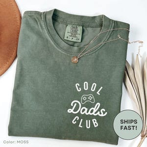 May include: A moss green Comfort Colors t-shirt with the text "COOL Dads CLUB" in white. The shirt has a crew neck and short sleeves. A gold necklace with a pendant is draped across the shirt. The shirt is size S-P-CH.