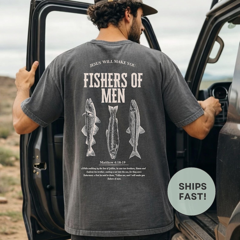 Puede incluir: Camiseta gris oscuro con el texto "JESUS WILL MAKE YOU FISHERS OF MEN" y tres ilustraciones de peces. El texto "Matthew 4:18-19" est&aacute; debajo de las ilustraciones de peces. La camiseta la lleva una persona junto a un veh&iacute;culo.