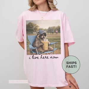 K&ouml;nnte beinhalten: Hellrosa T-Shirt mit einem Aufdruck eines Waschb&auml;ren mit Sonnenbrille und Hut, der in einem Stuhl sitzt und ein Getr&auml;nk trinkt. Der Text "i live here now" steht darunter. Das Shirt ist hellrosa.