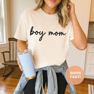 K&ouml;nnte beinhalten: Cremefarbenes T-Shirt mit dem Schriftzug "boy mom" in schwarzer Schrift. Die Person h&auml;lt einen hellblauen Becher mit goldenem Boden. Das Shirt wird mit einer schwarzen Hose und einer Jeansjacke kombiniert.