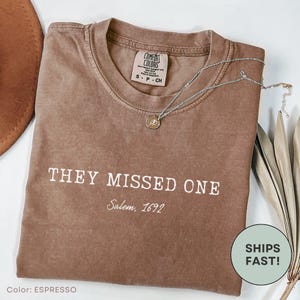 May include: A brown short-sleeved t-shirt featuring the white text "THEY MISSED ONE" and "Salem, 1692" printed on the front. The garment includes a Comfort Colors brand label at the inner neckline and is styled with a silver-toned rose pendant necklace.