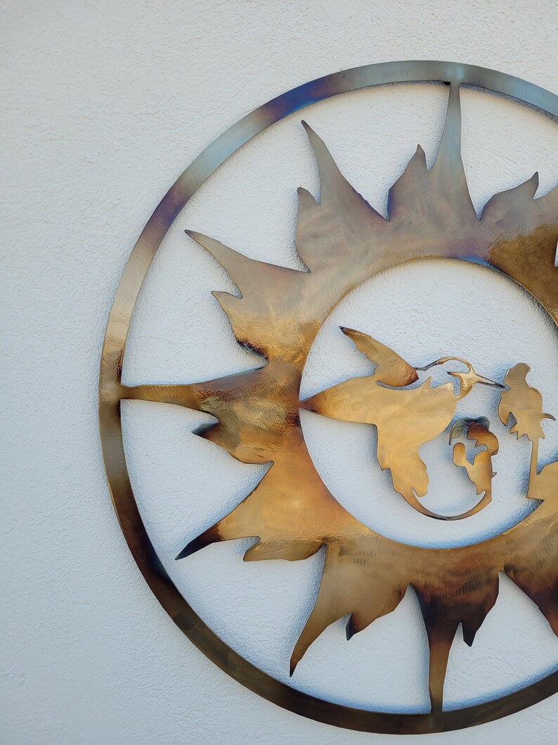 May include: A metal wall art piece in the shape of a sun with a hummingbird flying through it. The sun is a golden colour and the hummingbird is a lighter gold colour.