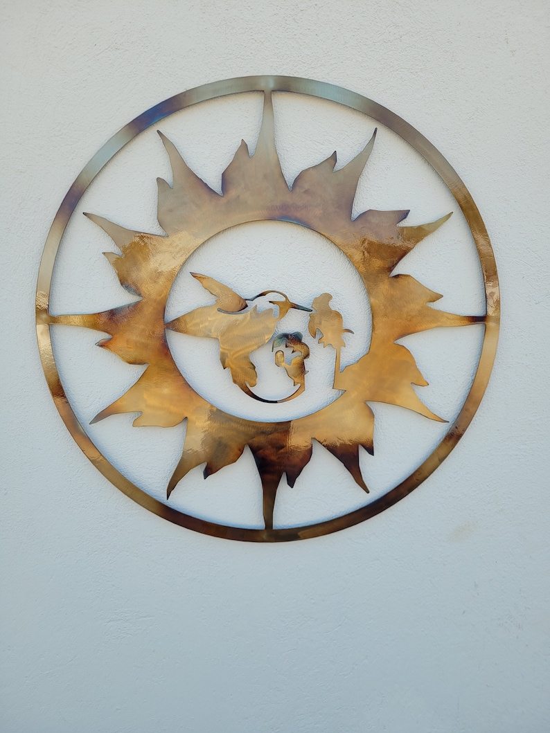 May include: A circular metal wall art piece with a sunburst design. The sunburst is made of metal and has a brown and gold finish. A hummingbird is silhouetted in the centre of the sunburst.