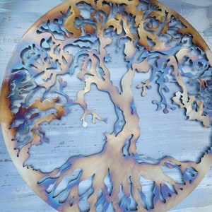 Op de afbeelding: Een cirkelvormig metalen wandkunstwerk dat een boom met ingewikkelde takken en wortels voorstelt. Het metaal heeft een gepatineerde afwerking met tinten bruin, goud en blauw.