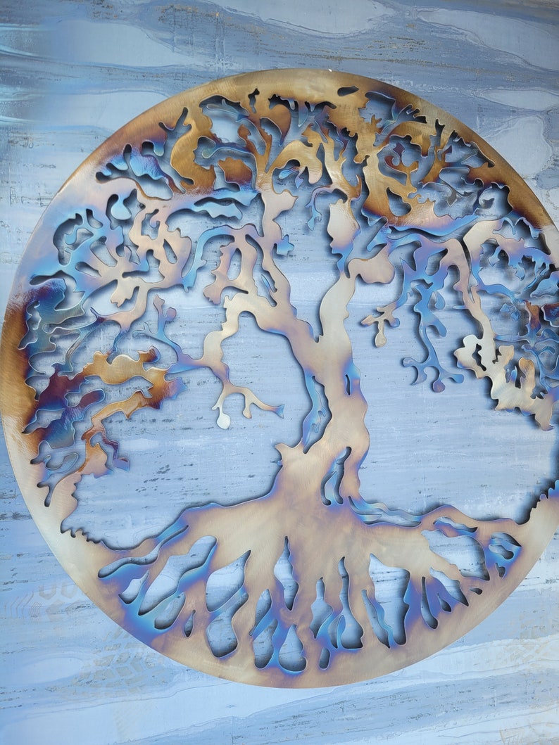 Op de afbeelding: Een cirkelvormig metalen wandkunstwerk dat een boom met ingewikkelde takken en wortels voorstelt. Het metaal heeft een gepatineerde afwerking met tinten bruin, goud en blauw.