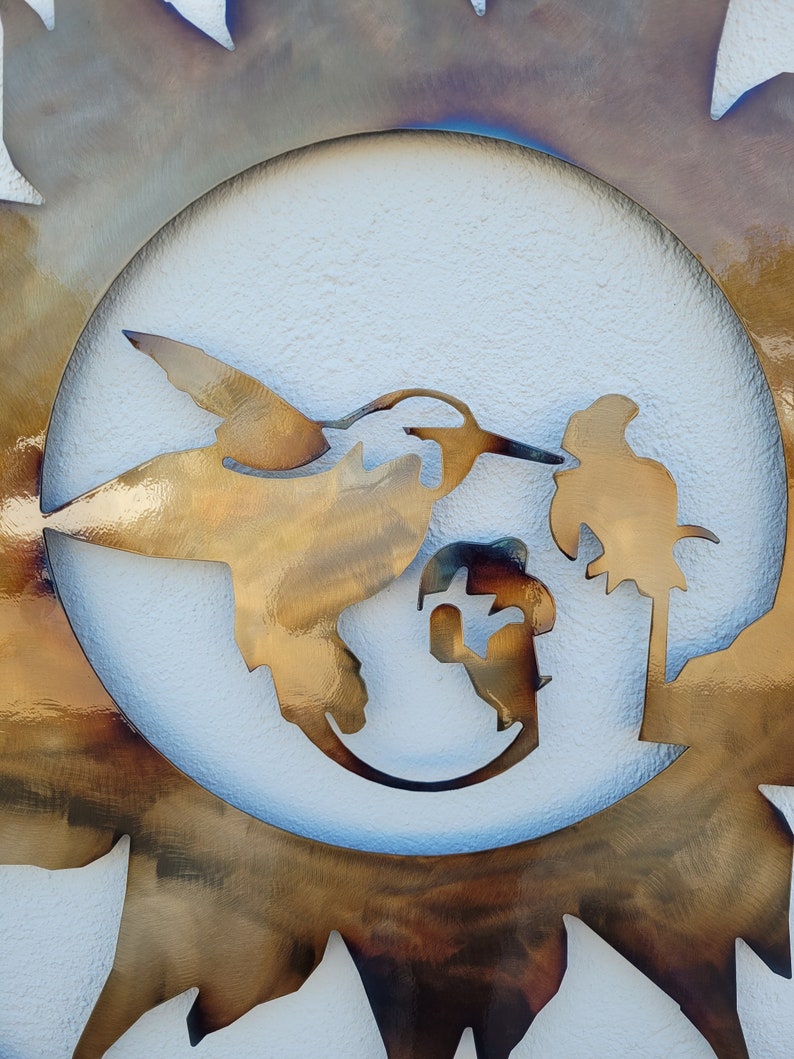 May include: A metal wall art piece depicting a hummingbird in flight with a flower and a bird perched on a branch. The art is set against a white background and surrounded by a sunburst design.