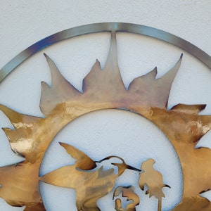 May include: A metal wall art piece featuring a stylised sun with a hummingbird and a globe inside. The sun is made of a gold-coloured metal and has a textured surface.