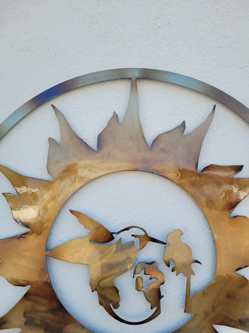 May include: A metal wall art piece featuring a stylised sun with a hummingbird and a globe inside. The sun is made of a gold-coloured metal and has a textured surface.