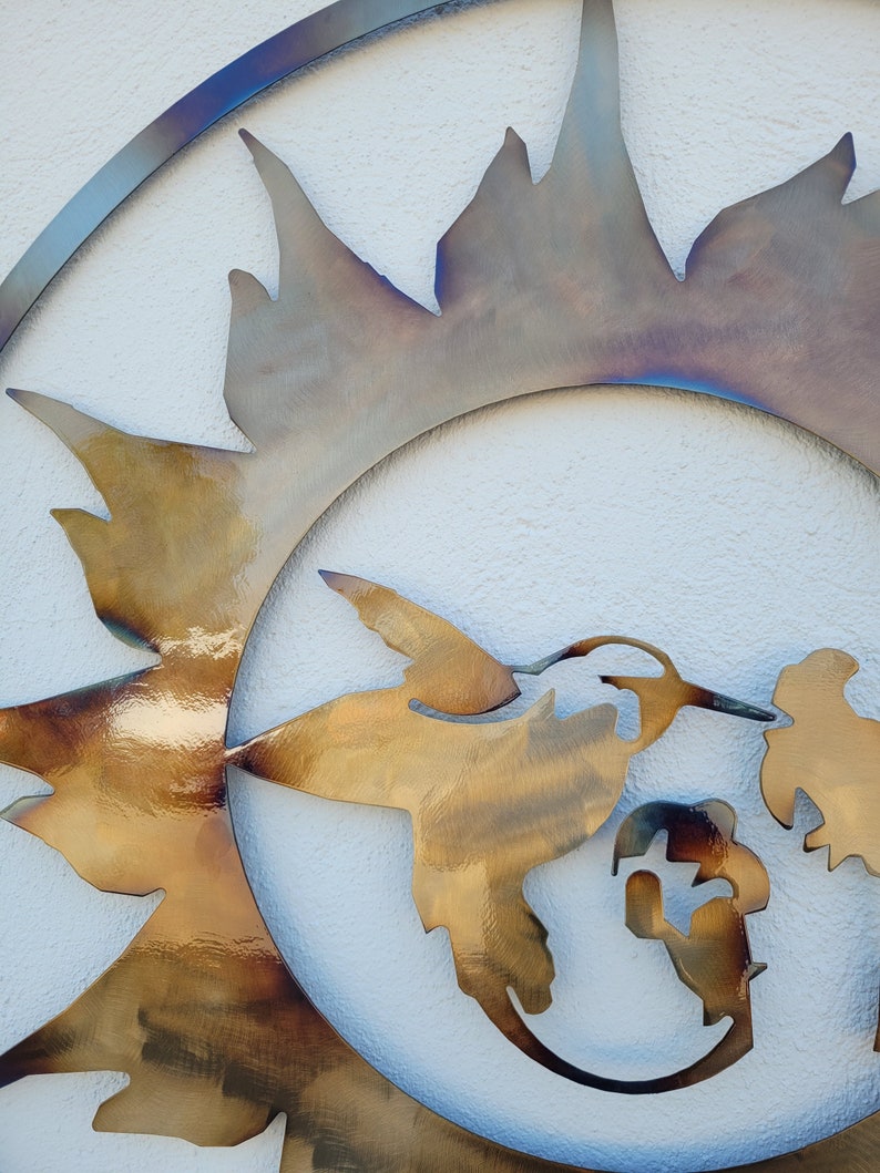 May include: A metal wall art sculpture featuring a hummingbird in flight, with a sunburst design behind it. The hummingbird is depicted in a golden colour, while the sunburst is a mix of gold and iridescent colours.