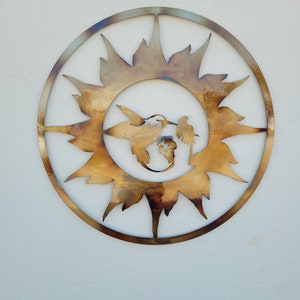 May include: A metal wall art piece in the shape of a sun with a hummingbird and a flower inside. The sun is made of a gold-coloured metal and has a rustic look.