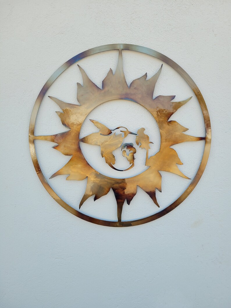 May include: A metal wall art piece in the shape of a sun with a hummingbird and a flower inside. The sun is made of a gold-coloured metal and has a rustic look.