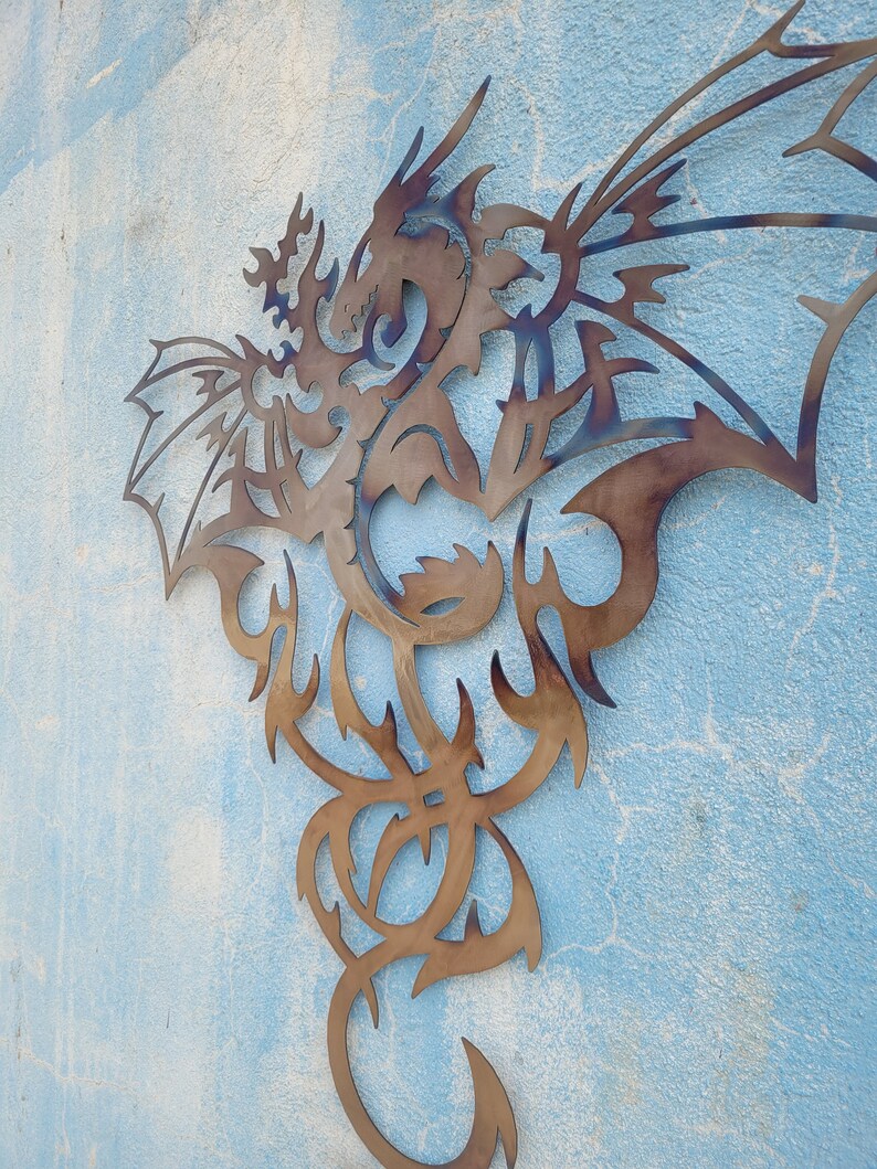 May include: A metal wall sculpture of a dragon with intricate details. The dragon is in a flying pose with its wings spread wide. The sculpture is made of a dark brown metal and has a rustic finish.