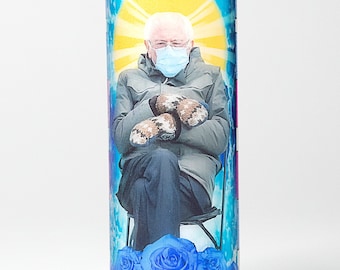 May include: A glass candle with a printed image of a man wearing a blue face mask and mittens, sitting in a chair with blue roses at the bottom. The background is a blue sky with a yellow sun.