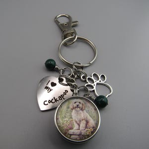 May include: A silver keychain with a heart charm that says "I love my Cockapoo" and a round charm with a picture of a white Cockapoo dog. The keychain also has a silver paw print charm and two green beads.