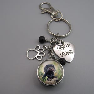 May include: A silver keychain with a charm featuring a black and white Cavapoo dog. The keychain also includes a silver paw print charm, a silver heart charm with the text "I love my Cavapoo", and black beads.