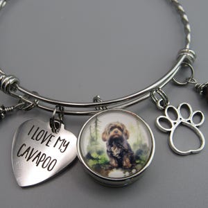 May include: A silver expandable bracelet with charms. The charms include a heart with the text "I love my Cavapoo", a paw print, a brown bead, and a round charm with a photo of a brown and white dog.