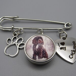 May include: Silver safety pin with three charms: a paw print, a round charm with a picture of a brown Cockapoo, and a heart-shaped charm with the text "I love my Cockapoo".