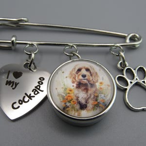 May include: A silver safety pin charm with three dangling charms. The first charm is a silver heart with the text "I love my Cockapoo". The second charm is a round glass charm with a picture of a brown and white Cockapoo dog. The third charm is a silver paw print.