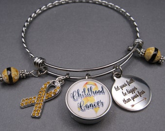 May include: A silver expandable bangle bracelet with three charms. The charms are a yellow ribbon with rhinestones, a round charm with the words "Childhood Cancer" and a yellow ribbon, and a round charm with the words "Let your faith be bigger than your fear" and an arrow.
