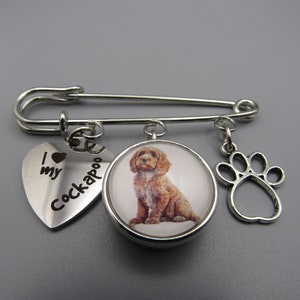 May include: Silver safety pin with three charms: a heart charm with the text "I love my Cockapoo", a round charm with a picture of a brown Cockapoo, and a paw print charm.