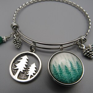 May include: A silver expandable bangle bracelet with multiple charms. The charms include two pine tree designs, one with a green and white glass cabochon and the other with a silver silhouette of two pine trees. The bracelet also has two green and white bead charms and two silver pine cone charms.
