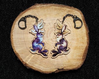 May include: Two acrylic keychains featuring jackalopes with antlers. One jackalope has a purple and blue galaxy-patterned body with light blue antlers, while the other has a brown and cream-colored body with tan antlers. Both have black metal rings.
