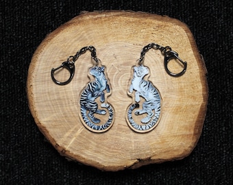 May include: Two acrylic keychains featuring a tiger illustration. One tiger is dark blue with black stripes, and the other is light blue with dark blue stripes. Both keychains have a black metal chain and a circular ring attachment.