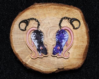 May include: Two acrylic keychains featuring rat illustrations with long pink tails and black metal rings. One rat has a black and white spotted coat, while the other displays a blue and purple galaxy pattern with white star-like speckles.