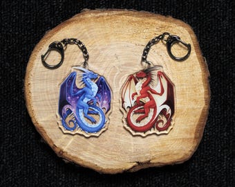 May include: Two acrylic keychains featuring dragon illustrations. One keychain displays a blue dragon with purple starry wings, while the other shows a red dragon with cream-colored wings. Both charms are attached to black metal key rings.