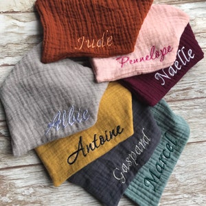 May include: A stack of six personalized muslin baby burp cloths in various colors, including gray, brown, pink, yellow, and green. The cloths are embroidered with the names Allie, Antoine, Gaspard, Marcel, Penelope, and Jude.