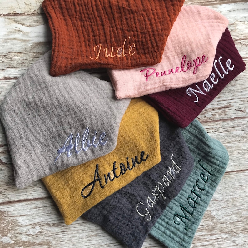 May include: A stack of six personalized muslin baby burp cloths in various colors, including gray, brown, pink, yellow, and green. The cloths are embroidered with the names Allie, Antoine, Gaspard, Marcel, Penelope, and Jude.