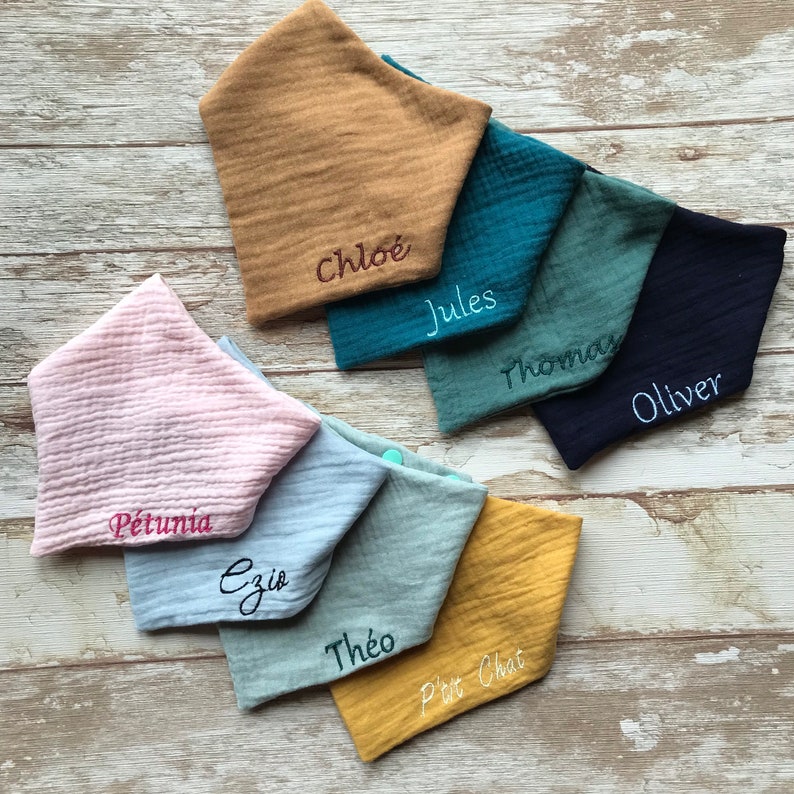 May include: Nine different colored fabric baby bibs with embroidered names on each. The names are: Chloe, Jules, Thomas, Oliver, Petunia, Ezio, Theo, and Petit Chat.