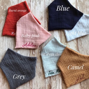 May include: A set of ten fabric baby bibs in various colors, including burnt orange, baby pink, light blue, blue, grey, vert d'eau clair, and camel. Each bib has a name embroidered on it.