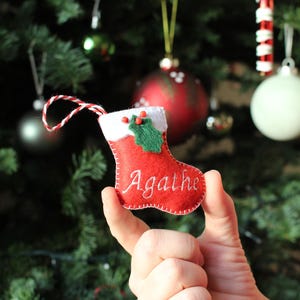 Op de afbeelding: Een kleine, rode kerstsok-ornament van vilt, geborduurd met de naam "Agathe" in het wit. Het heeft een witte manchet, groene hulstdetails en een rood-wit touwtje om op te hangen. Het ornament wordt in een hand gehouden.