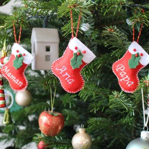 Puede incluir: Un árbol de Navidad decorado con calcetines de fieltro rojo y blanco con los nombres "Maman", "Aria" y "Papa" bordados en ellos. El árbol también está decorado con adornos, incluyendo una manzana roja y un adorno a rayas verdes y blancas.