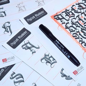 May include: A black Artline Supreme Calligraphy Pen 4.0 rests on a white surface scattered with calligraphy practice sheets. The sheets feature stylized blackletter uppercase letters and the text "Stylized Blackletter UPPERCASE WORKBOOK". A sample of calligraphy is in the upper right corner.