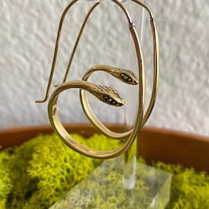 Könnte beinhalten: Goldfarbene Creolen mit Schlangen-Design. Jeder Ohrring hat einen gewundenen Schlangenkopf mit strukturierten Details und kleinen schwarzen Akzenten. Die Ohrringe werden auf einem transparenten Ständer vor grünem Moos präsentiert.