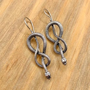 Pode incluir: Um par de brincos de cobra prateados. Cada brinco apresenta um design detalhado de cobra, com o corpo formando um símbolo de infinito e uma cabeça texturizada. Os brincos pendem de um gancho simples.