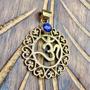May include: A gold-toned metal pendant featuring a circular frame with intricate scrollwork patterns surrounding a central Om symbol. A small, round, deep blue stone is set at the top of the pendant, just below the metal bail for a necklace chain.