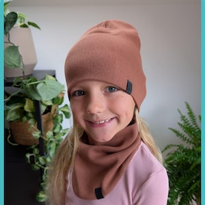 May include: A brown ribbed knit beanie and neck gaiter set. The beanie and neck gaiter have a small black tag on the side. The child is wearing a pink long-sleeved shirt.