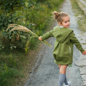 May include: Two children, a girl and a boy, walk hand-in-hand along a dirt track. The girl wears a green hooded jacket, grey socks, and white trainers. The boy wears a dark blue jacket, grey trousers, and orange and black trainers. Both hold tall grass.