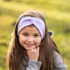 May include: A young girl with long brown hair is wearing a light purple headband and a grey hooded sweatshirt. She is smiling and looking at the camera.