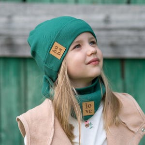 Może przedstawiać: Mała dziewczynka w zielonym wełnianym berecie i dopasowanym kominie. Berecik ma brązową skórzaną naszywkę z napisem "bu ba". Komin ma brązową skórzaną naszywkę z napisem "love".
