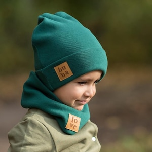 May include: A teal blue knitted hat and matching scarf with brown leather patches that say "buba" and "love".